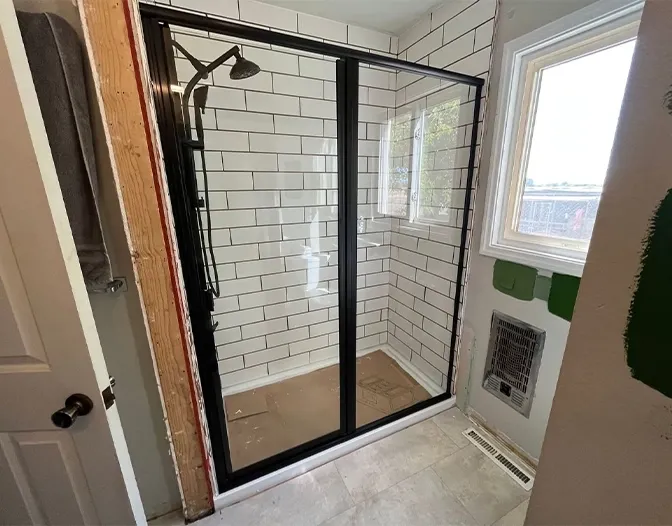 A newly installed shower with white subway tile walls, black-framed sliding glass doors, and a ceiling-mounted showerhead in a bathroom under renovation.