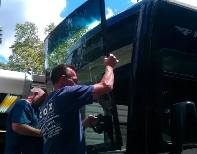 Two workers are installing a large windshield on a commercial vehicle using suction tools, with trees and a blue sky in the background.