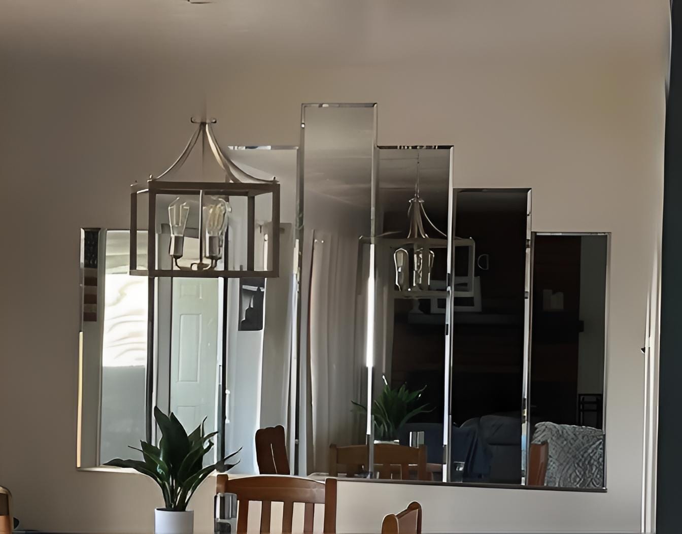 Custom beveled mirrors and a modern chandelier are reflected on a beige wall above a wooden dining table with a small potted plant.