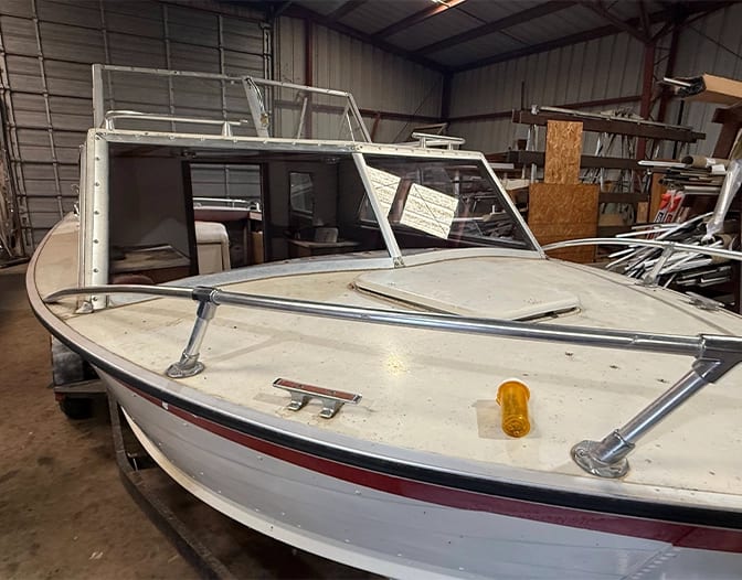 A white boat with a red stripe is parked inside a metal storage building. The boat’s windshield is partially open, resembling a glass panel shower enclosure, and a small yellow object rests on the deck.