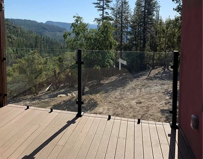 A wooden deck with a sleek deck glass railing overlooks a dry, wooded landscape with trees and hills in the background.