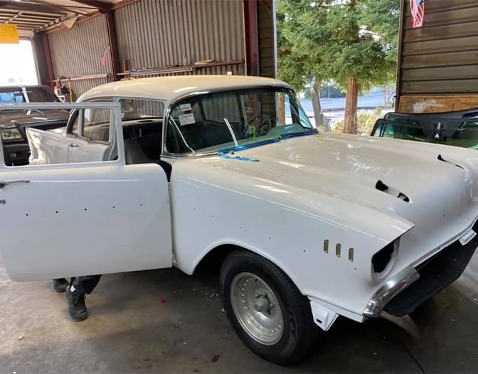 A classic white car with its driver-side door open is parked inside a garage; parts of its hood and trim appear unfinished.