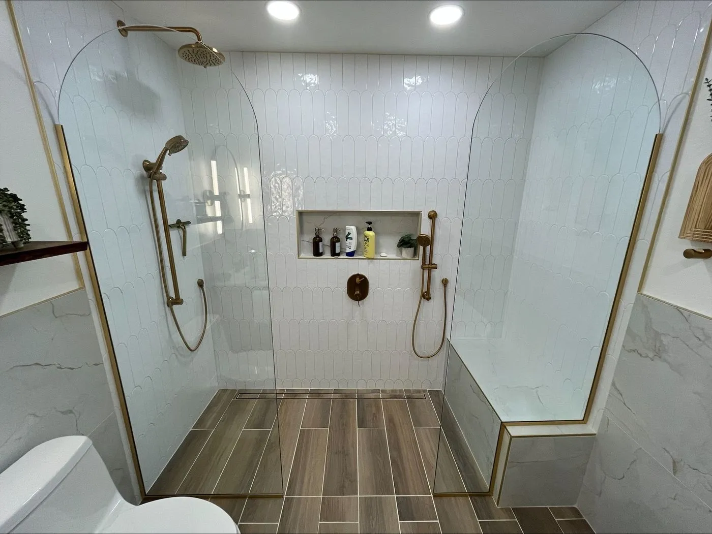 Modern bathroom with a double shower setup, glass dividers, gold fixtures, a built-in bench, niche shelves, and wood-look tile flooring.