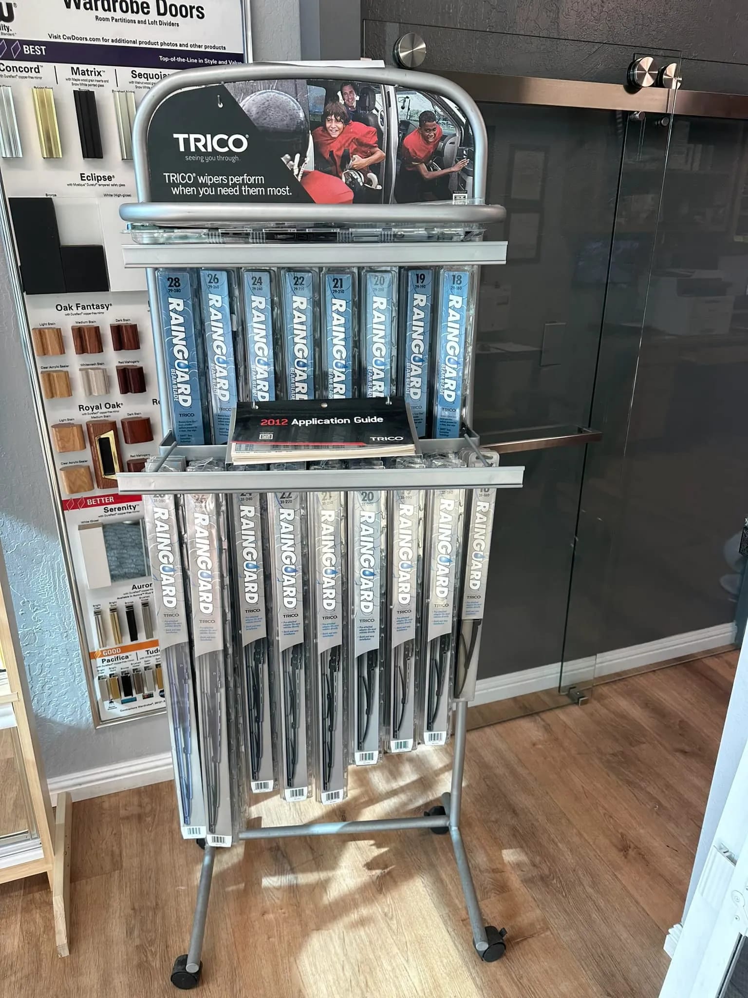 A display stand with multiple TRICO RainGuard windshield wiper blades and an application guide, positioned on a wooden floor near a glass door.