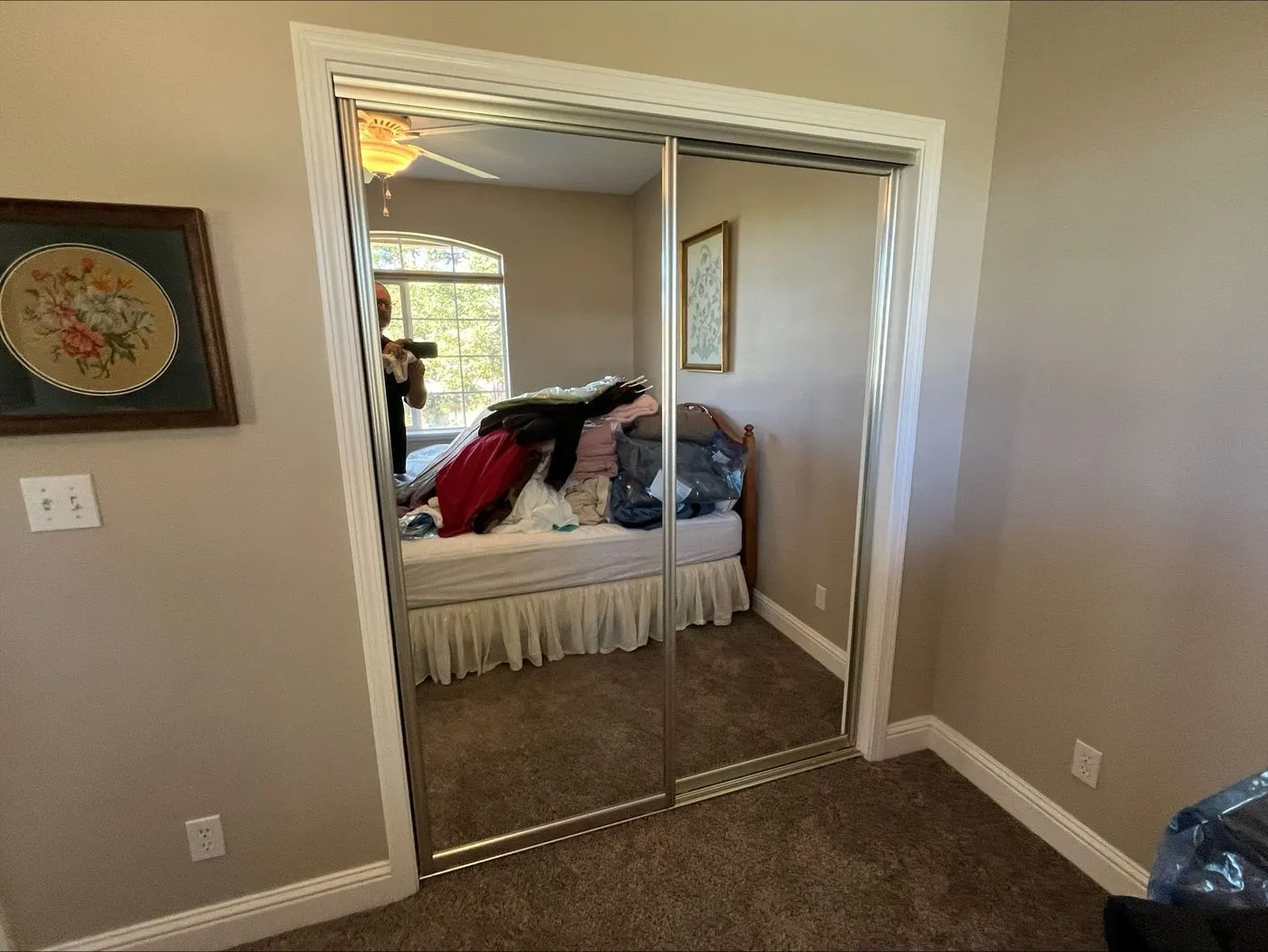 A bedroom with mirrored closet doors reflects a bed covered in clothes, beige walls, framed art, and brown carpet. A person is partially visible taking the photo.