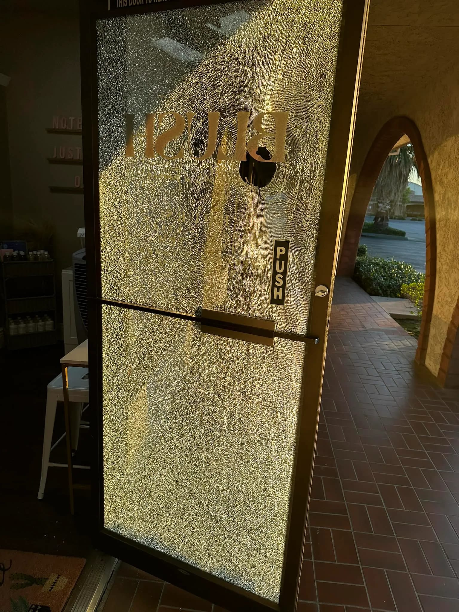 A glass door with a shattered pane and a hole near the top, with sunlight casting shadows on the door. The word "PUSH" is visible on the door’s handle.
