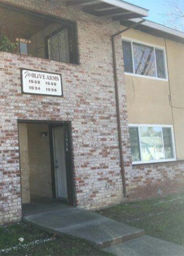 A two-story brick and stucco apartment building with a sign reading "The Olive Arms" and a concrete walkway leading to the entrance.