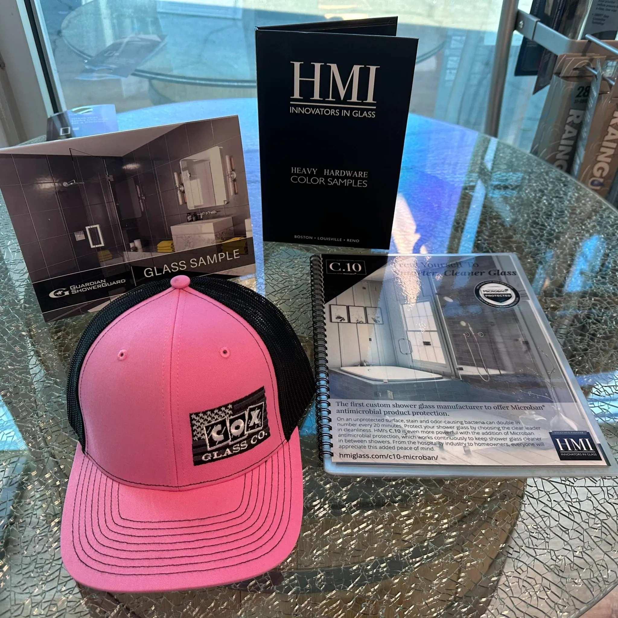 A pink and black COX Glass Co. hat sits on a glass table with glass sample booklets and an HMI Innovators in Glass brochure displayed behind it.