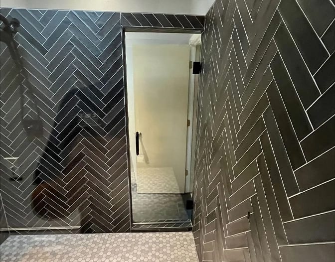 A modern bathroom with black herringbone tile walls, a frameless glass shower door enclosure, and a floor with small hexagonal tiles.