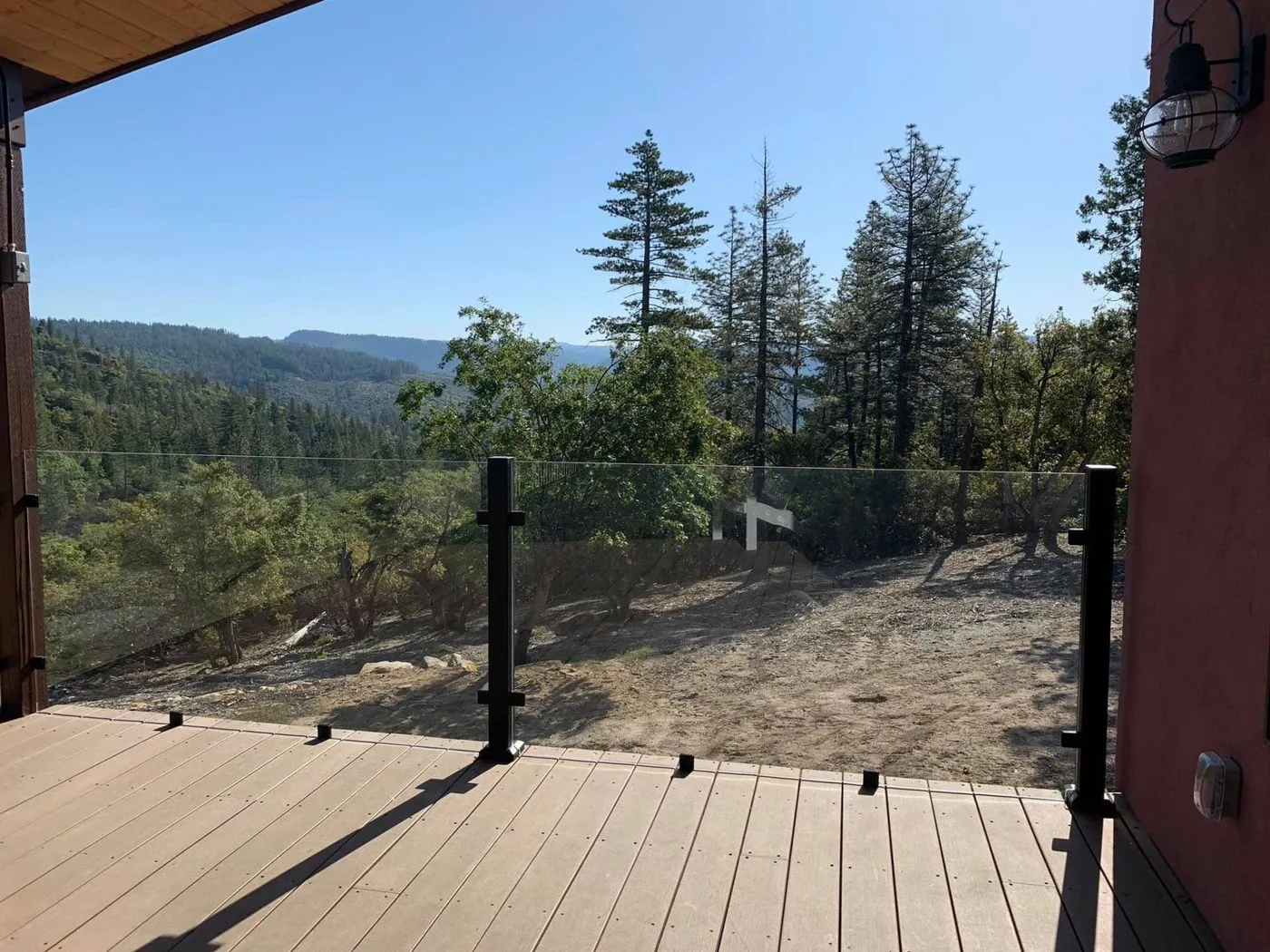 A wooden deck with a glass railing overlooks a dirt clearing and a forested mountain landscape under a clear blue sky.