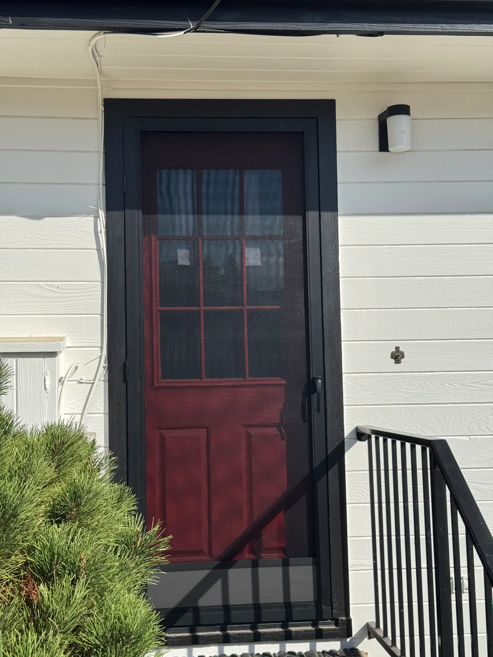 A red screen door with black trim on a white house, next to a small outdoor light and a metal railing, with greenery in the lower left corner.