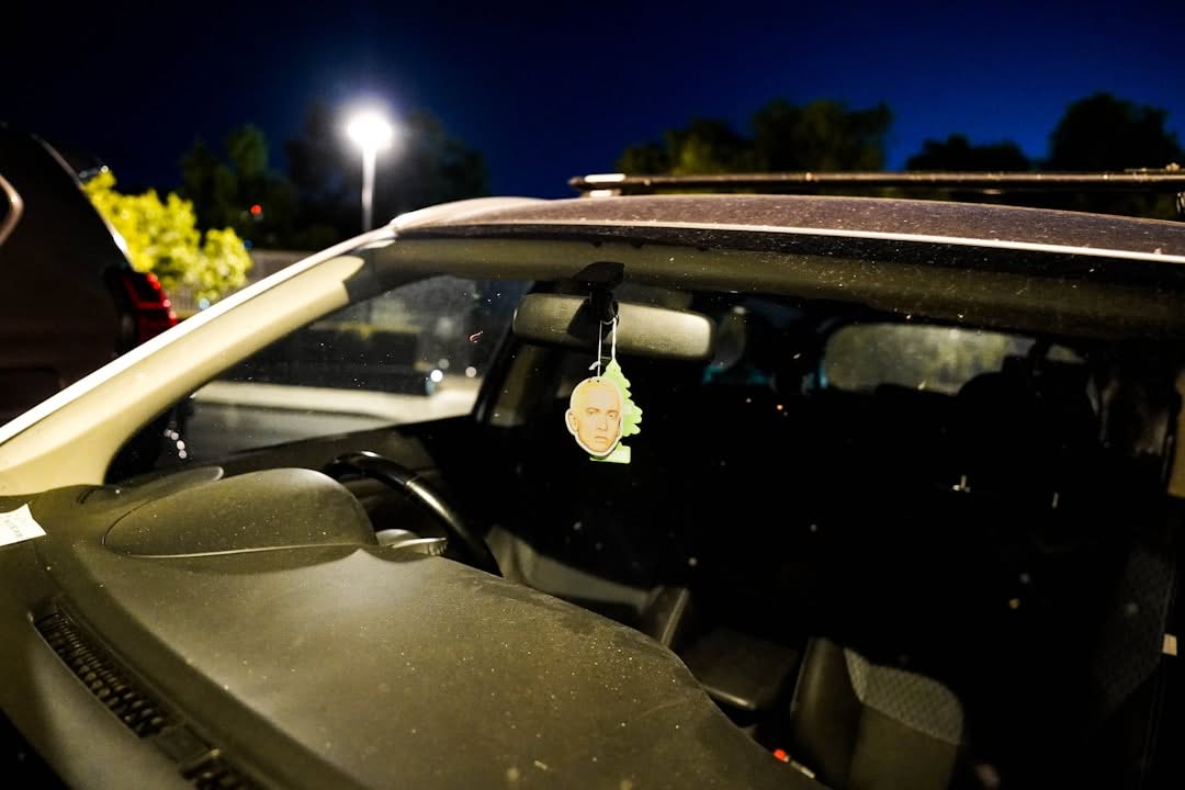 A car interior at night with a cartoon face air freshener hanging from the rearview mirror; the windshield is dusty and a streetlamp is visible outside.