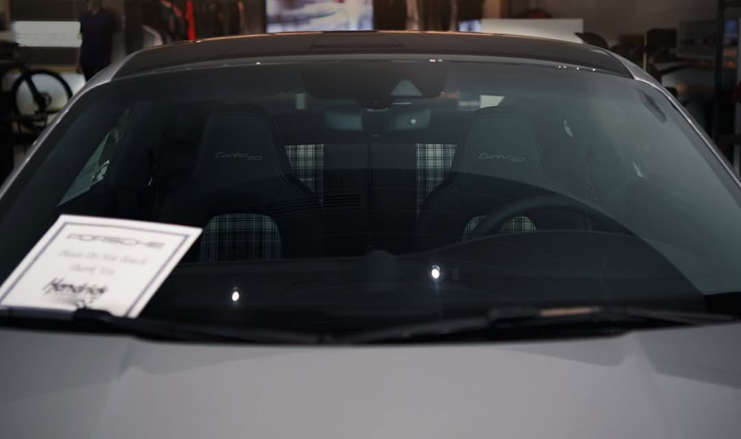 View through the windshield of a parked Porsche, showing two plaid-patterned front seats and a dealership sign on the dashboard.