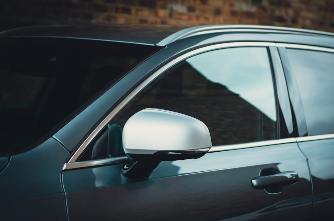 Close-up of the driver side door, window, and side mirror of a modern dark-colored car parked in front of a brick wall.