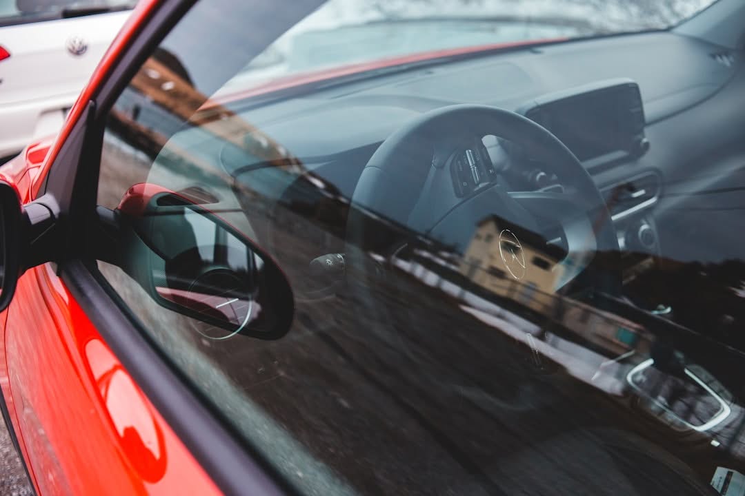 Close-up view through the window of a red car, showing the steering wheel, dashboard, and driver's side mirror. Reflections on the glass are visible.