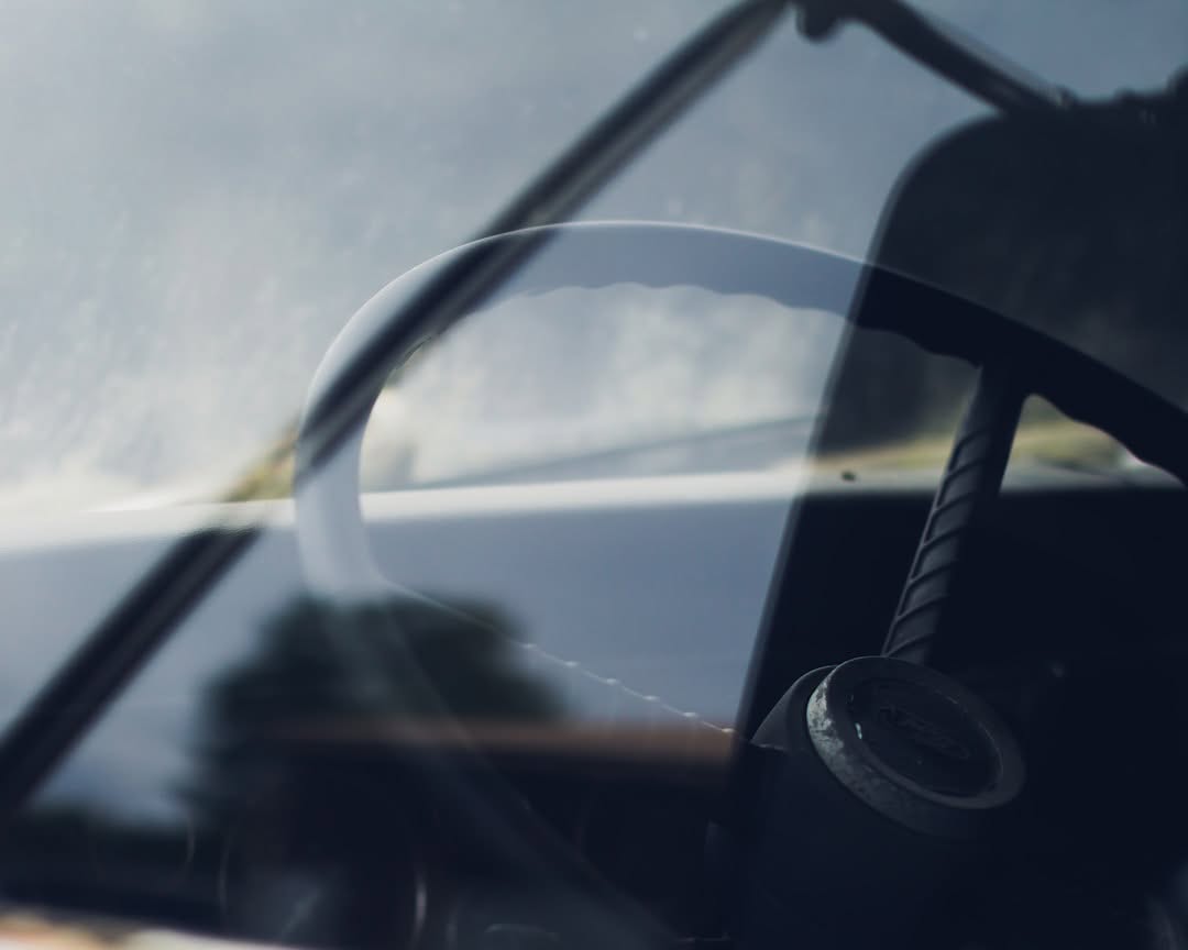 Close-up view of a vintage car steering wheel and dashboard, seen through the windshield with reflections visible on the glass.