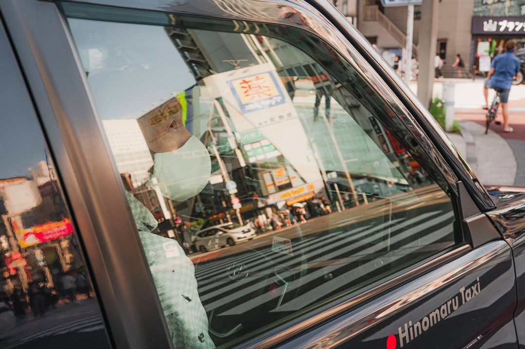 A person wearing a mask sits inside a Hinomaru taxi, visible through the car window, with a busy city street and crosswalk reflected in the glass.