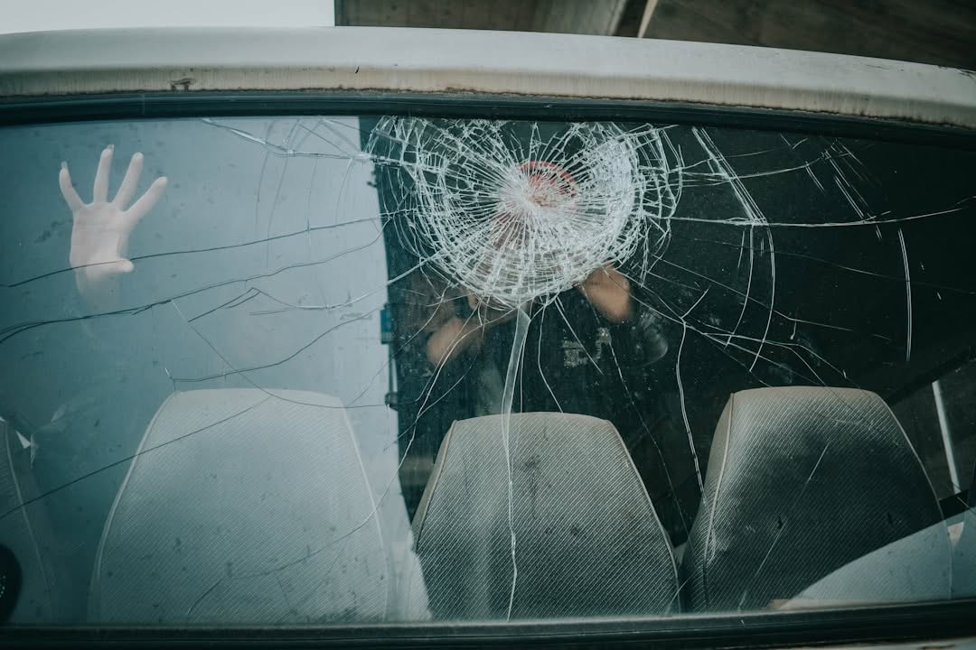 A person’s hand is pressed against a car window with a large, circular crack in the glass; two empty seats are visible inside.