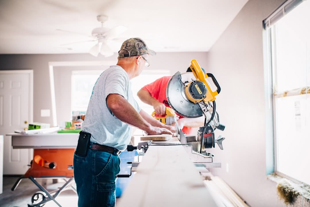 Two men are working indoors with a circular saw, cutting wooden planks on a long workbench near a window.