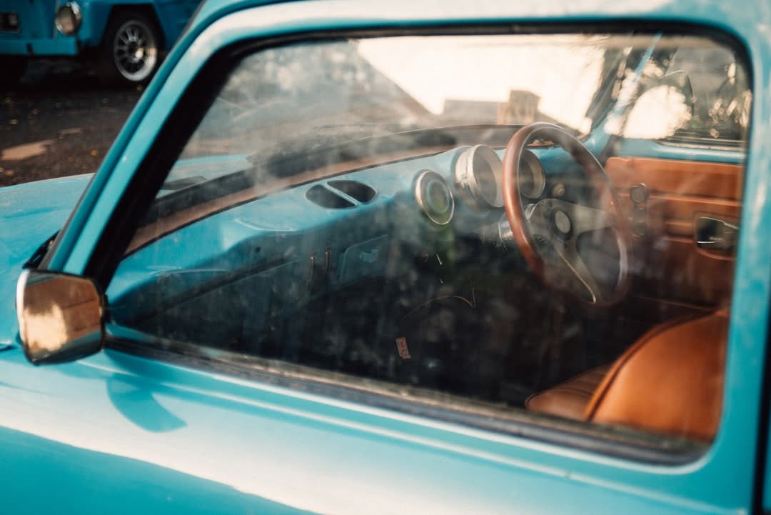 Close-up view through the window of a vintage blue car, showing the steering wheel, dashboard, and brown seat upholstery.