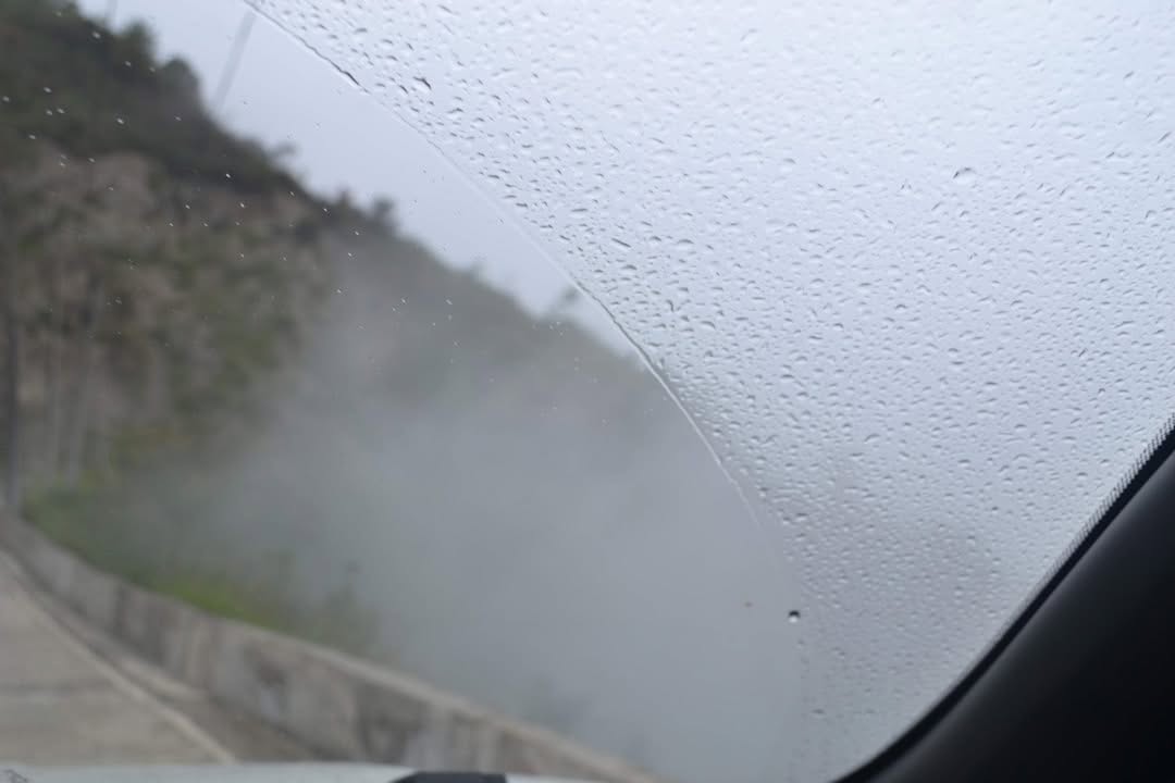 View through a car windshield with raindrops and fog, showing a misty road curving along a hillside.