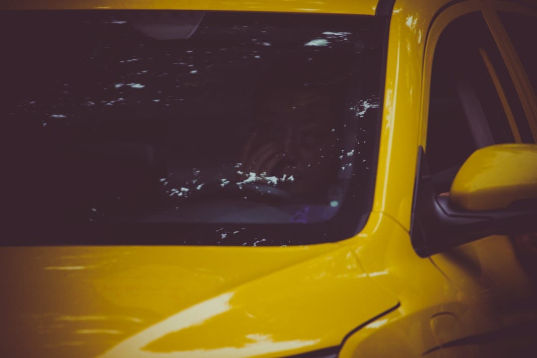 A person sits inside a yellow car, partially visible through a windshield with reflections and smudges.