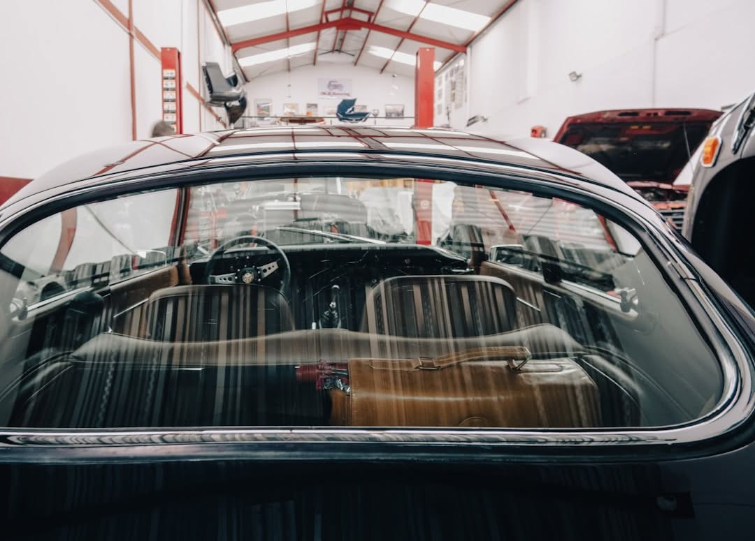 View through the rear window of a classic car inside an auto repair shop, showing the interior seats, steering wheel, and surrounding workshop equipment.