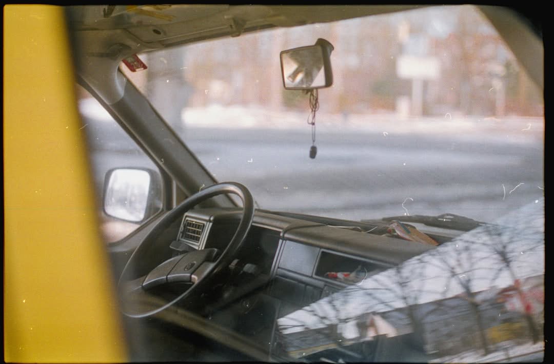 View through a car window showing the steering wheel, dashboard, and a small mirror hanging from the ceiling. Reflections of trees and sky are visible on the glass.