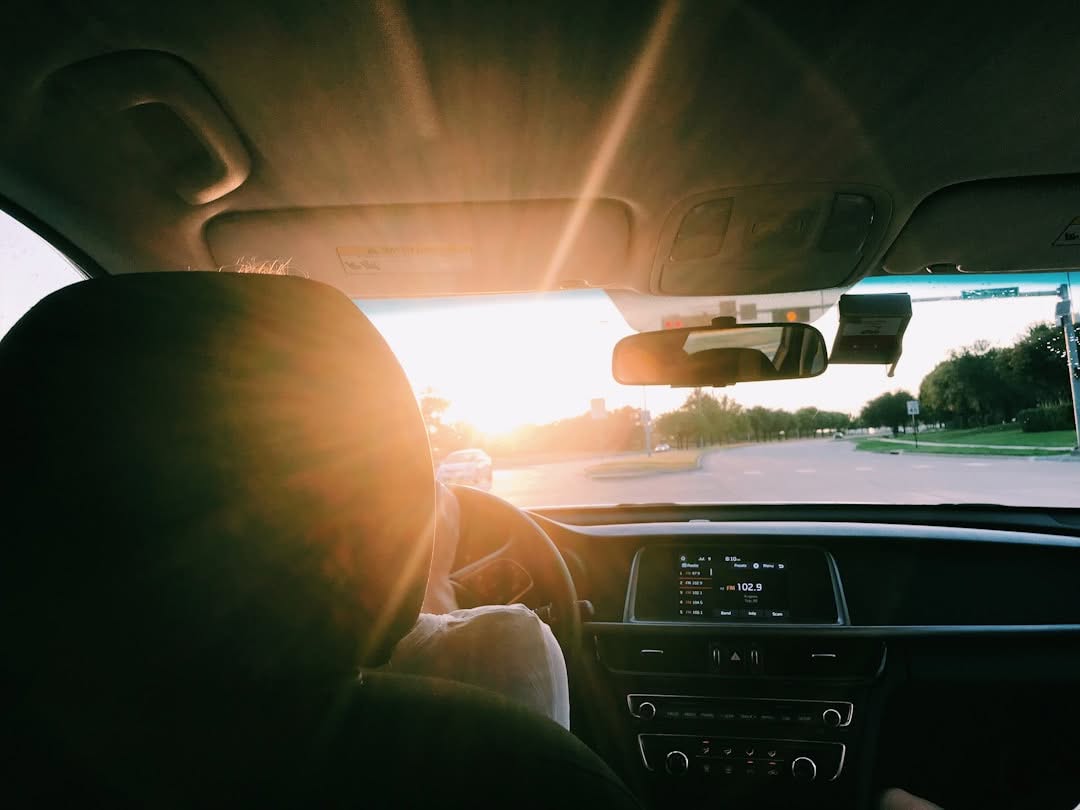 View from inside a car at sunset, showing a driver at the wheel and sunlight shining through the windshield onto the dashboard and road ahead.