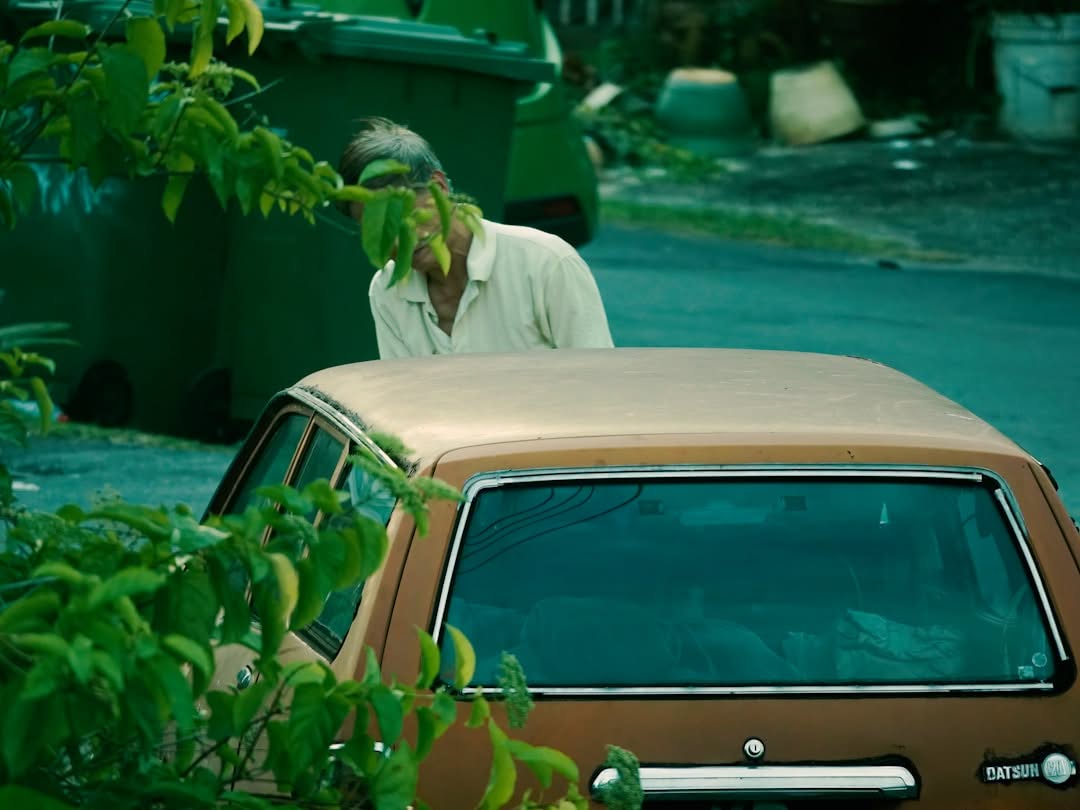 An older man in a light shirt stands next to a vintage brown Datsun car parked by the roadside, partially obscured by green leaves.